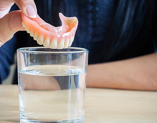 Someone gently placing their dentures into a glass of clear fluid with two fingers