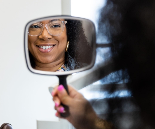 Woman with glasses admiring her smile in a mirror after visiting dentist in Jenks