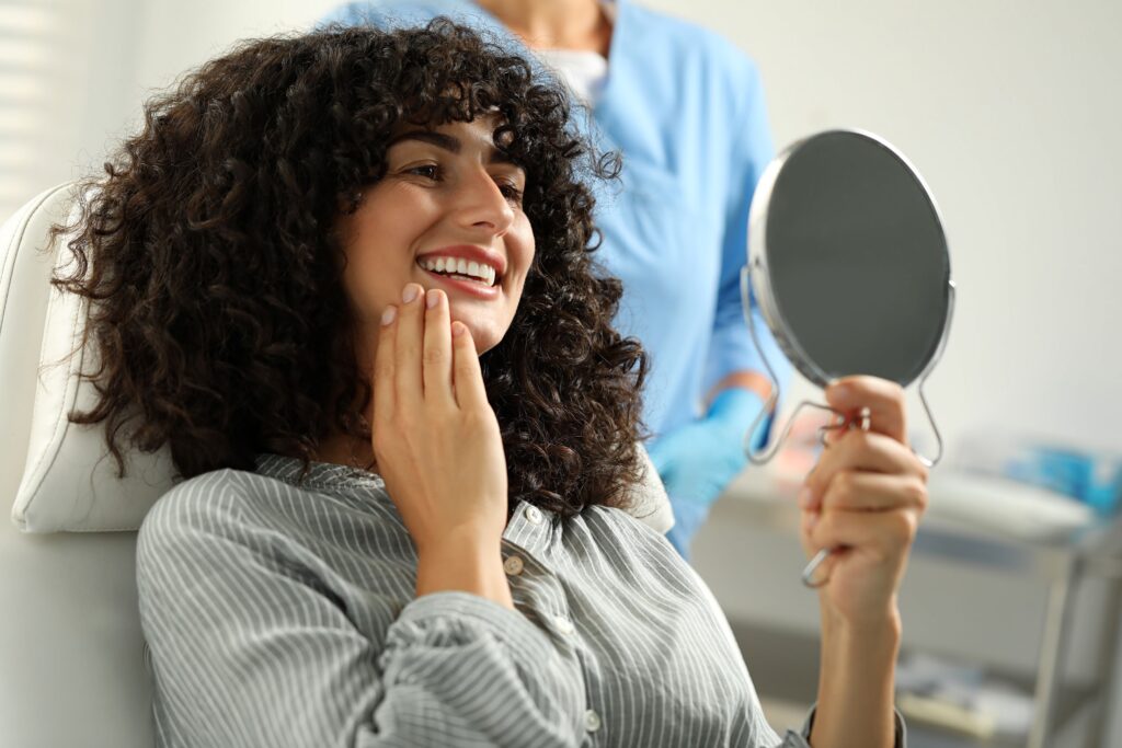 Woman with dark curly hair in dental chair smiling at her reflection